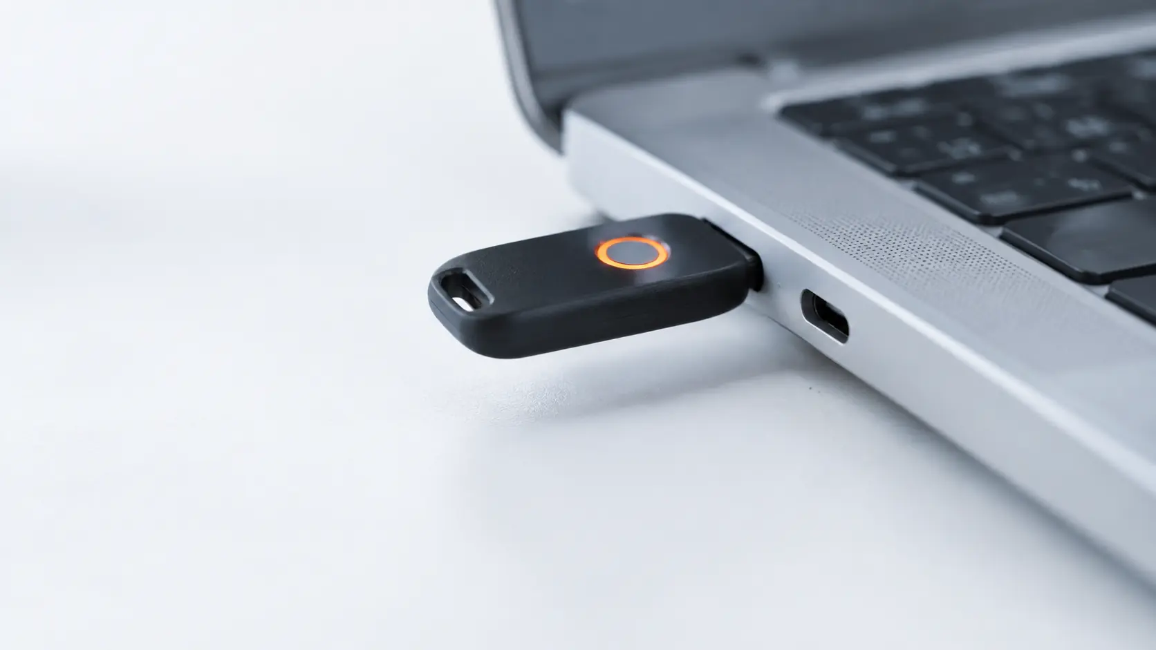 A small hardware security key plugged into the side of a laptop, its touch sensor glowing orange.